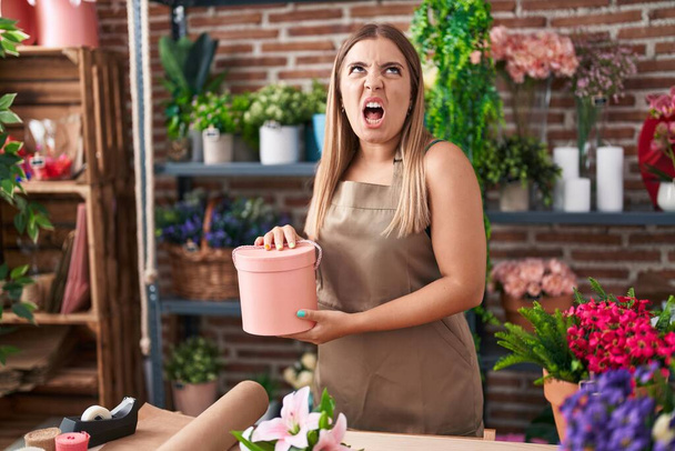 jonge blonde vrouw werken bij bloemist winkel boos en gek schreeuwen gefrustreerd en furieus, schreeuwen van woede op zoek omhoog.  - Foto, afbeelding