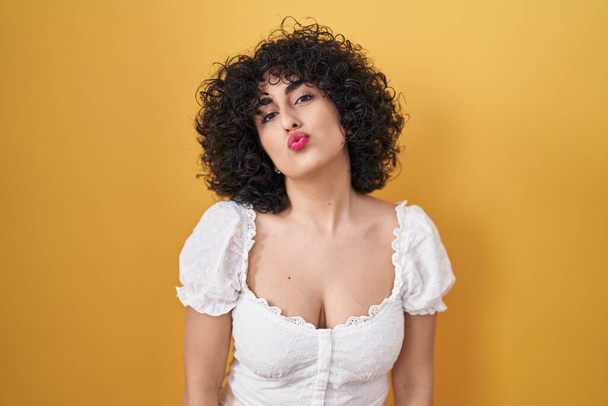 Young brunette woman with curly hair standing over yellow background looking at the camera blowing a kiss on air being lovely and sexy. love expression.  - Photo, Image