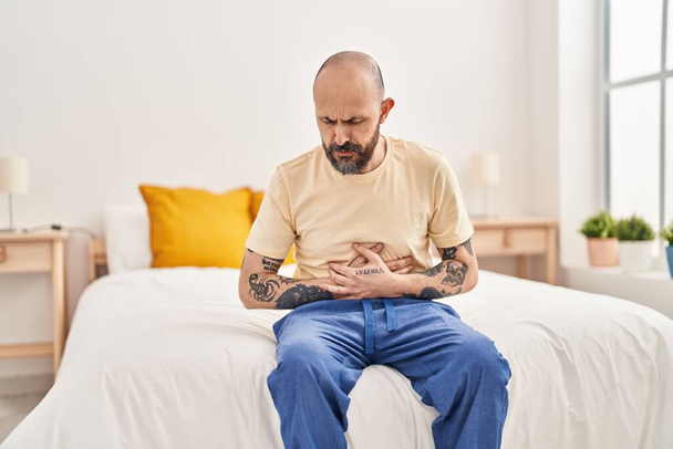 Молодой лысый мужчина страдает от боли в животе, сидя на кровати в спальне - Фото, изображение