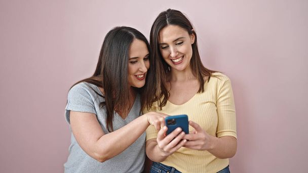 Dwie kobiety uśmiechające się pewnie za pomocą smartfona na odizolowanym różowym tle - Zdjęcie, obraz