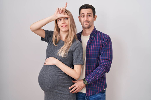 Young couple expecting a baby standing over white background making fun of people with fingers on forehead doing loser gesture mocking and insulting.  - Photo, Image