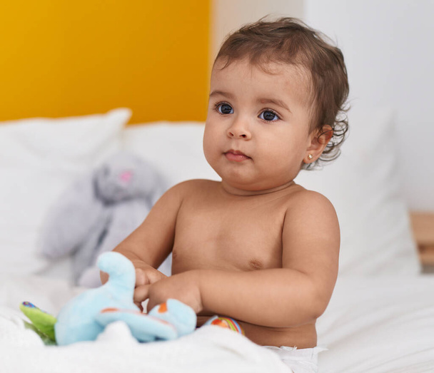 Adorável bebê hispânico sentado na cama brincando com boneca elefante no quarto - Foto, Imagem