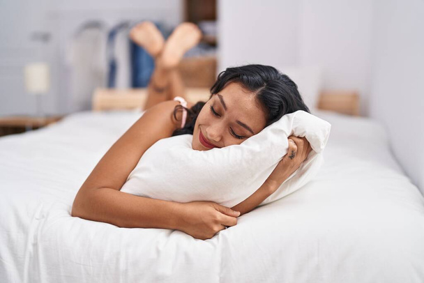 Jovem bela mulher hispânica abraçando travesseiro deitado na cama no quarto - Foto, Imagem