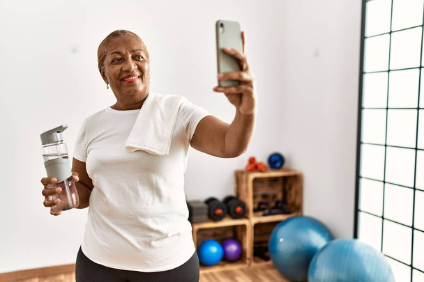Старша афро-американська жінка, посміхаючись впевнено робить вибір за допомогою смартфона в спортивному центрі. - Фото, зображення