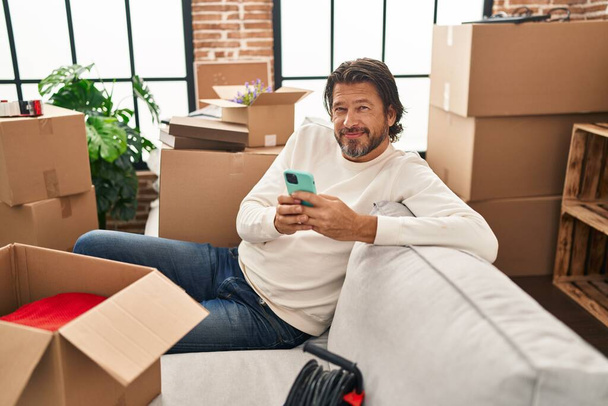 Mann mittleren Alters sitzt mit Smartphone auf Sofa im neuen Zuhause - Foto, Bild