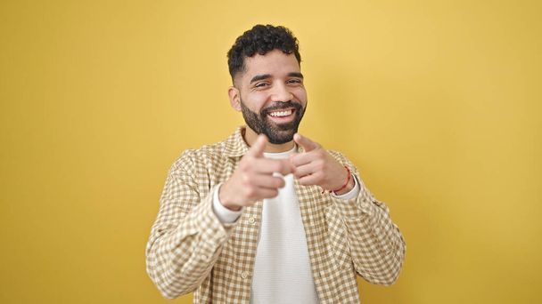 Fiatal spanyol férfi mosolygós magabiztos mutató ujjal a kamera felett elszigetelt sárga háttér - Fotó, kép