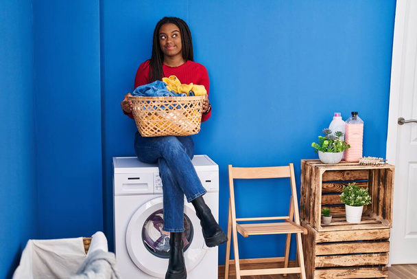 Junger Afroamerikaner mit Zöpfen und Wäschekorb auf Waschmaschine sitzend, lächelnd zur Seite blickend und nachdenklich wegstarrend.  - Foto, Bild