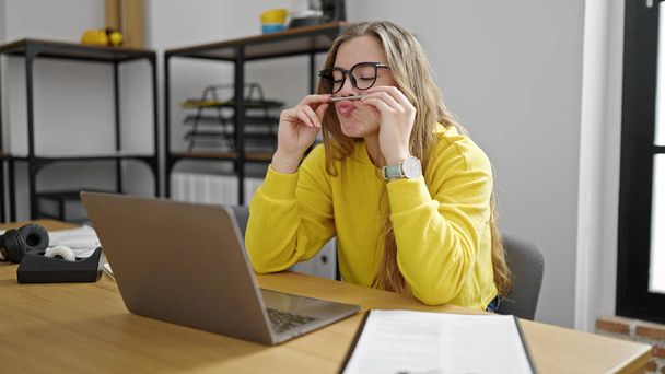 Jeune femme blonde femme d'affaires faisant un geste drôle avec un stylo sur la bouche au bureau - Photo, image