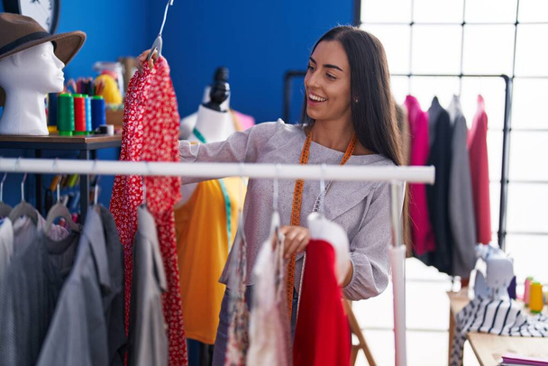 Jeune belle femme hispanique tailleur souriant confiant tenant des vêtements sur rack à l'usine de vêtements - Photo, image