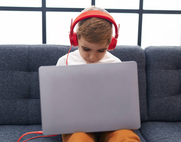 Adorable hispanic boy using laptop and headphones sitting on sofa at home - Zdjęcie, obraz