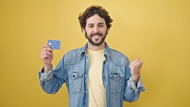 Mladý hispánec drží kreditní kartu s vítězným výrazem nad izolovaným žlutým pozadím - Fotografie, Obrázek