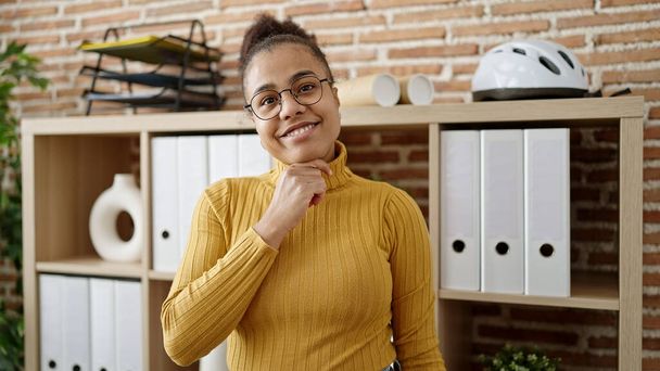 Nuori afrikkalainen amerikkalainen nainen hymyilee luottavainen yllään lasit toimistossa - Valokuva, kuva