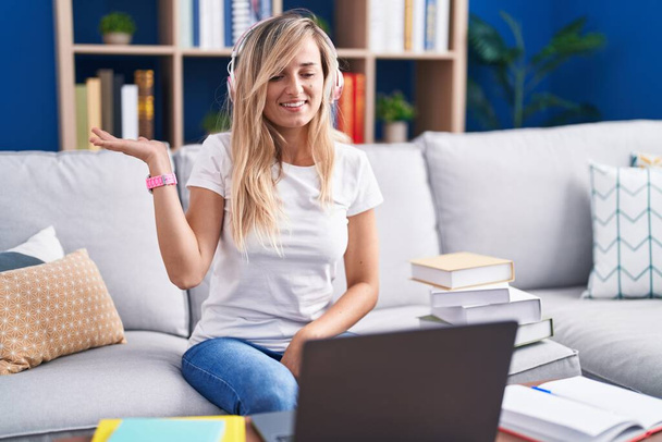 Młoda blondynka studiuje za pomocą laptopa komputerowego w domu uśmiechnięta, radosna prezentacja i wskazująca dłonią na aparat fotograficzny.  - Zdjęcie, obraz