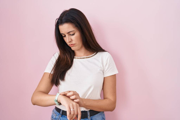 Young brunette woman standing over pink background checking the time on wrist watch, relaxed and confident  - Foto, afbeelding