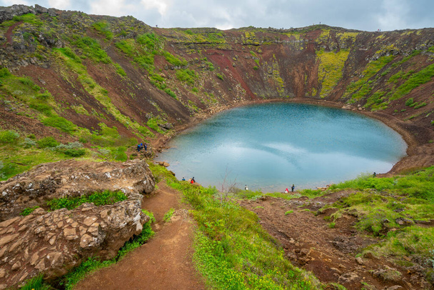 Draufsicht auf den Kerid-Kratersee in Island an einem bewölkten Tag mit Menschen, die in der Ferne spazieren gehen und herumstehen. Türkisfarbenes Wasser der isländischen Kraterlagune - Foto, Bild