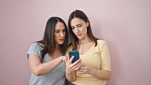 Dos mujeres usando smartphone con expresión seria sobre fondo rosa aislado - Foto, Imagen