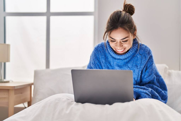 Молода красива іспаномовна жінка використовує ноутбук, сидячи на ліжку в спальні
 - Фото, зображення