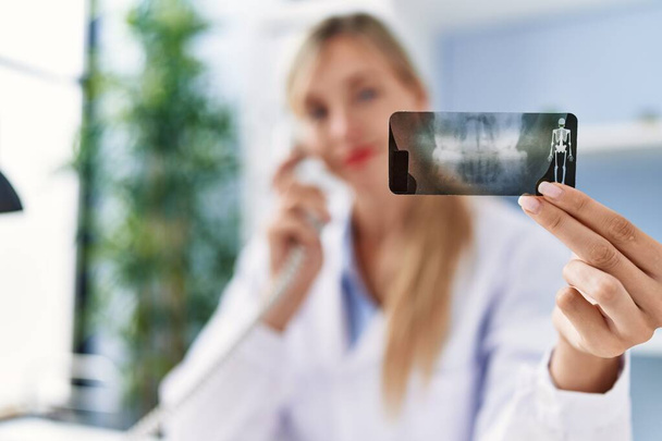 Молодая блондинка в форме дантиста разговаривает по телефону, держа рентген в клинике - Фото, изображение