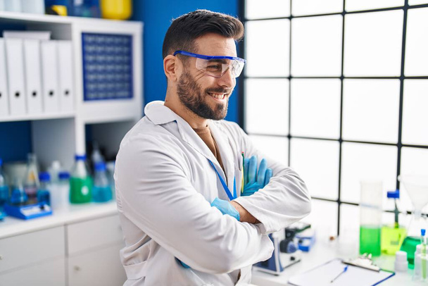 Joven hispano vistiendo uniforme científico sentado con los brazos cruzados gesto en el laboratorio - Foto, imagen
