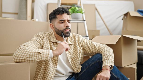 Young hispanic man sitting on floor with doubt expression at new home - Foto, imagen