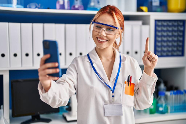 Junge kaukasische Frau, die im wissenschaftlichen Labor arbeitet und Videotelefonie mit Smartphone macht, lächelnd mit einer Idee oder Frage, Zeigefinger mit glücklichem Gesicht, Nummer eins  - Foto, Bild