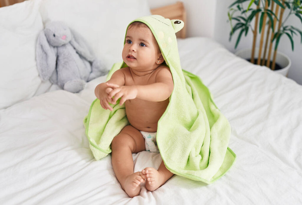 Adorabile bambino ispanico che indossa un asciugamano di rana seduto sul letto in camera da letto - Foto, immagini