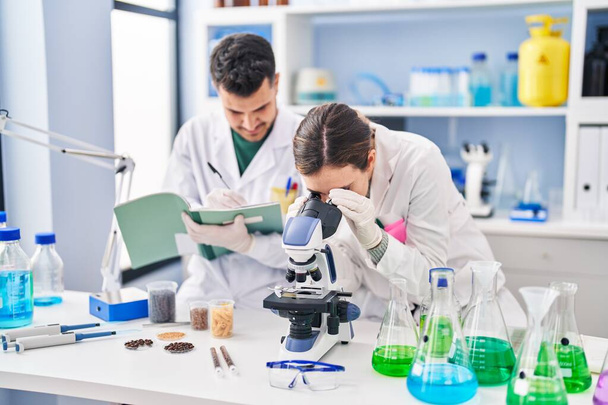 Hombre y mujer vistiendo uniforme científico usando escritura de microscopio en notebook en laboratorio - Foto, imagen
