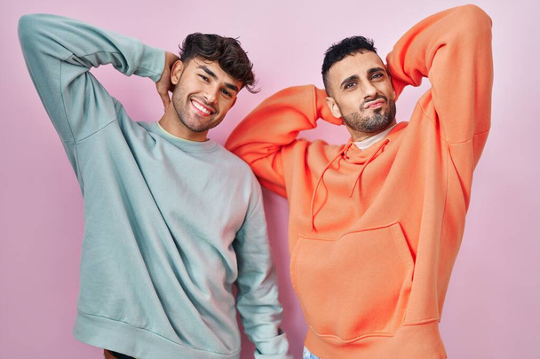 Joven pareja gay hispana de pie sobre fondo rosa relajante y estiramiento, brazos y manos detrás de la cabeza y el cuello sonriendo feliz  - Foto, Imagen