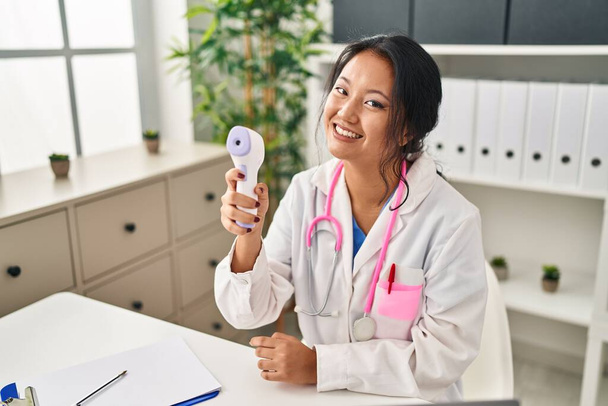Young chinese woman wearing doctor uniform using thermometer at clinic - Foto, Imagen