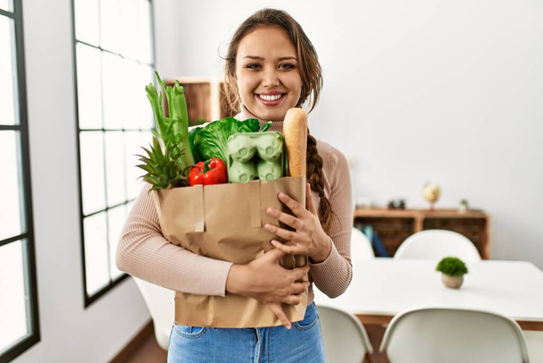 Joven hermosa mujer hispana sonriendo confiada sosteniendo bolsa de comestibles en casa - Foto, Imagen