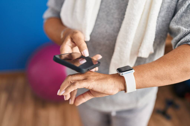 Middle age hispanic woman wearing sportswear using smartwatch and smartphone at laundry room - Foto, imagen