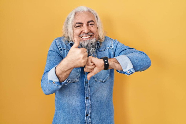 Middle age man with grey hair standing over yellow background doing thumbs up and down, disagreement and agreement expression. crazy conflict  - Foto, afbeelding