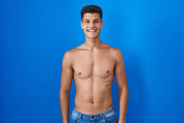 Mladý hispánec stojící bez košile nad modrým pozadím se šťastným a chladným úsměvem na tváři. šťastný člověk.  - Fotografie, Obrázek