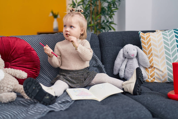 Liebenswerte blonde Mädchen Vorschulkind sitzt auf Sofa Zeichnung auf Notizbuch zu Hause - Foto, Bild