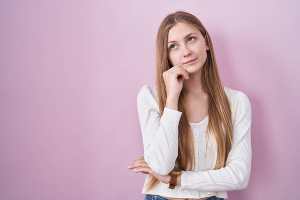 Young caucasian woman standing over pink background with hand on chin thinking about question, pensive expression. smiling with thoughtful face. doubt concept.  - Fotografie, Obrázek