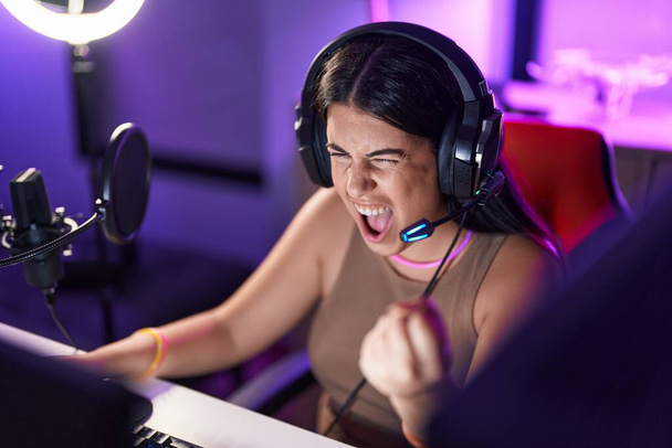 Jeune belle hispanique femme streamer jouer jeu vidéo avec l'expression gagnante à la salle de jeux - Photo, image