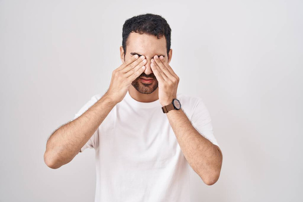 Hombre hispano guapo parado sobre fondo blanco frotando ojos para fatiga y dolor de cabeza, expresión somnolienta y cansada. problema de visión  - Foto, imagen