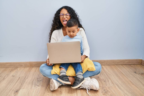 Jeune mère hispanique et enfant utilisant ordinateur portable assis sur le sol langue collante heureux avec expression drôle. concept d'émotion.  - Photo, image