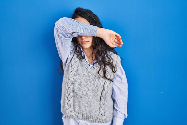Молодая брюнетка женщина стоит на синем фоне закрывая глаза с рукой, глядя серьезно и грустно. беспросветная, скрывающая и отвергающая концепция  - Фото, изображение