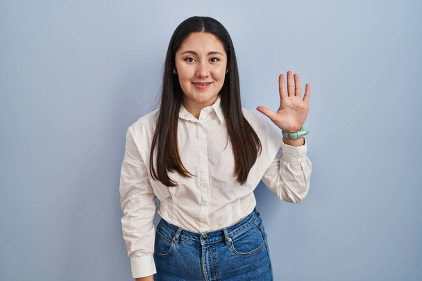 Young latin woman standing over blue background showing and pointing up with fingers number five while smiling confident and happy.  - Zdjęcie, obraz