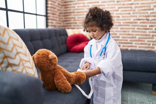 Adorável hispânico menina vestindo médico uniforme examinando ursinho de pelúcia em casa - Foto, Imagem