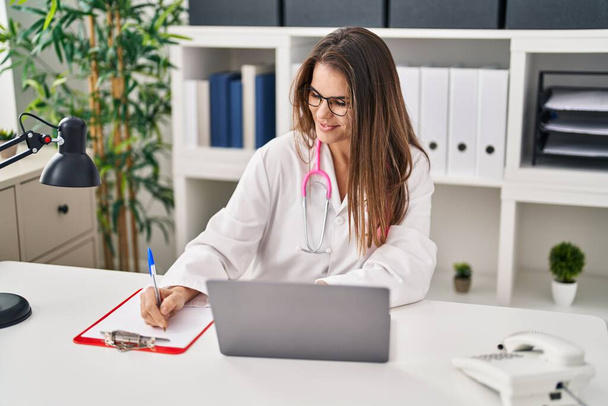 Young beautiful hispanic woman doctor using laptop writing on clipboard at clinic - Фото, изображение