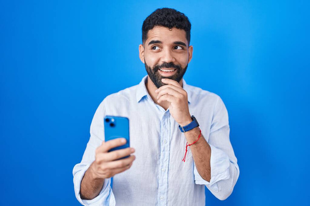 Hispanic man with beard using smartphone typing message with hand on chin thinking about question, pensive expression. smiling and thoughtful face. doubt concept.  - Zdjęcie, obraz