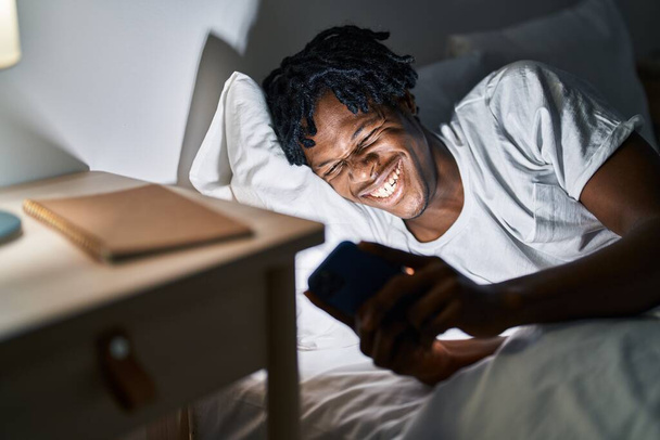 Αφροαμερικάνος που χρησιμοποιεί smartphone ξαπλωμένο στο κρεβάτι στο υπνοδωμάτιο - Φωτογραφία, εικόνα