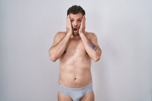 Jeune homme hispanique debout torse nu portant des sous-vêtements effrayés et choqués, surprise et expression étonnante avec les mains sur le visage  - Photo, image