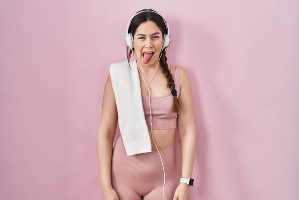 スポーツウェアとヘッドフォンを身に着けている若いブルネットの女性は面白い表情で幸せ舌を突き出す。感情の概念.  - 写真・画像