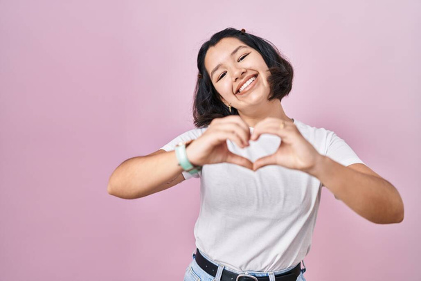 Latin kökenli genç bir kadın pembe arka planda gelişigüzel beyaz tişörtler giyiyor. Sevgiyle gülümsüyor. Elleriyle kalp sembolü yapıyor. romantik kavram.  - Fotoğraf, Görsel