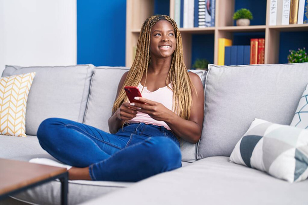 Afrikkalainen amerikkalainen nainen käyttää älypuhelinta istuu sohvalla kotona - Valokuva, kuva
