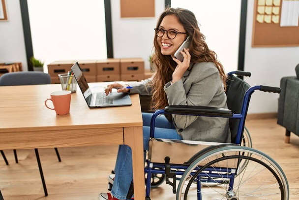 Junge schöne hispanische Geschäftsfrau spricht im Büro auf dem Smartphone sitzend im Rollstuhl - Foto, Bild