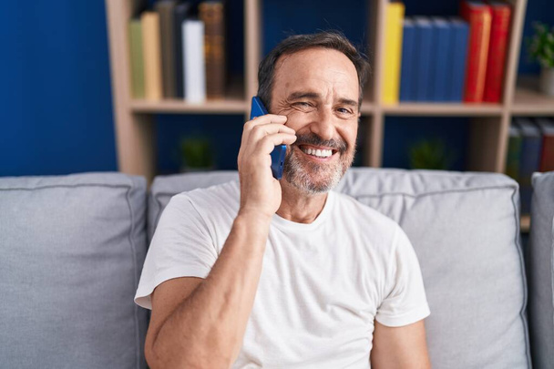 Мужчина средних лет разговаривает по смартфону, сидя дома на диване - Фото, изображение